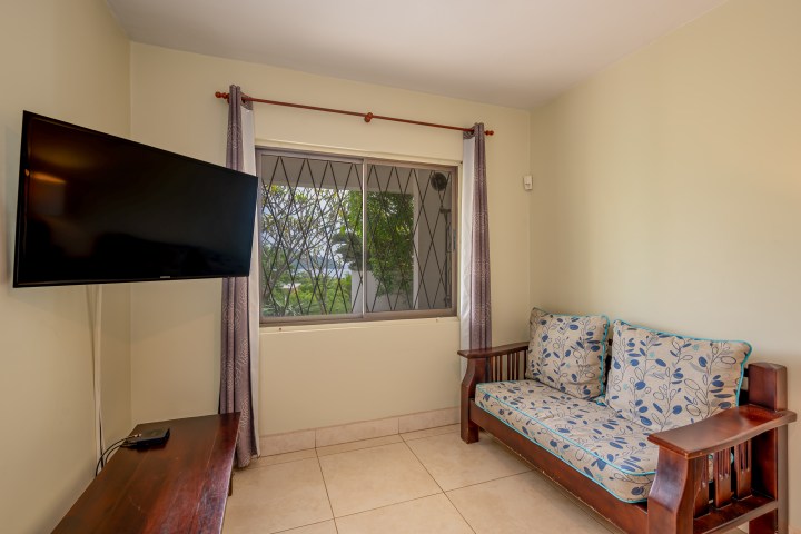 Room with a wall-mounted TV, wooden sofa with floral cushions, and a window with bars and curtains.