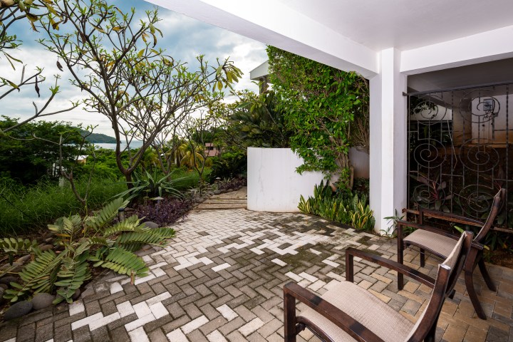 Patio with chairs, surrounded by lush greenery and brick flooring, overlooking distant trees.