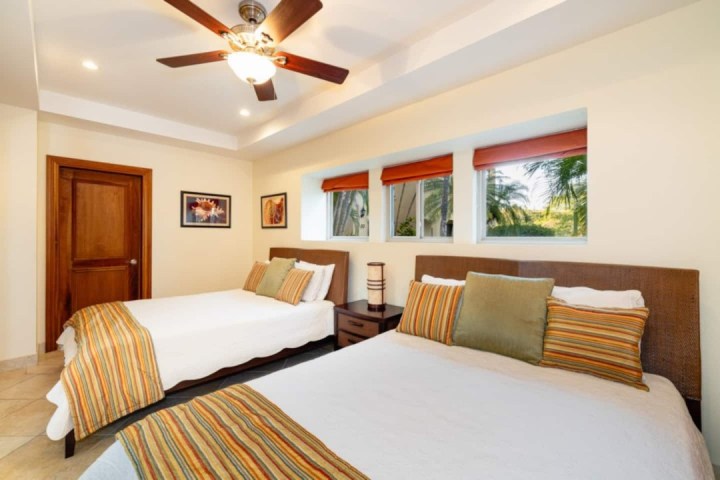 Bedroom with two beds, striped pillows, ceiling fan, and windows with palm tree view.