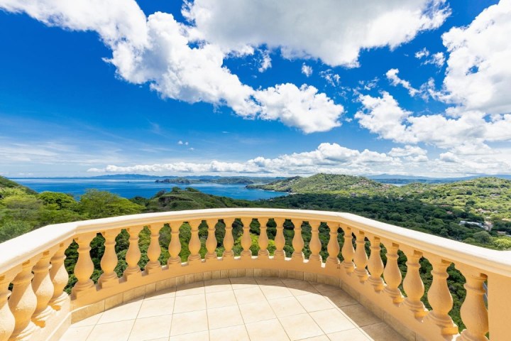 Balcony with stone railing overlooking lush hills and a distant ocean under a bright blue sky with clouds.