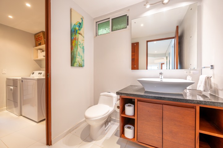 Modern bathroom with a sink, toilet, large mirror, and a small laundry area.