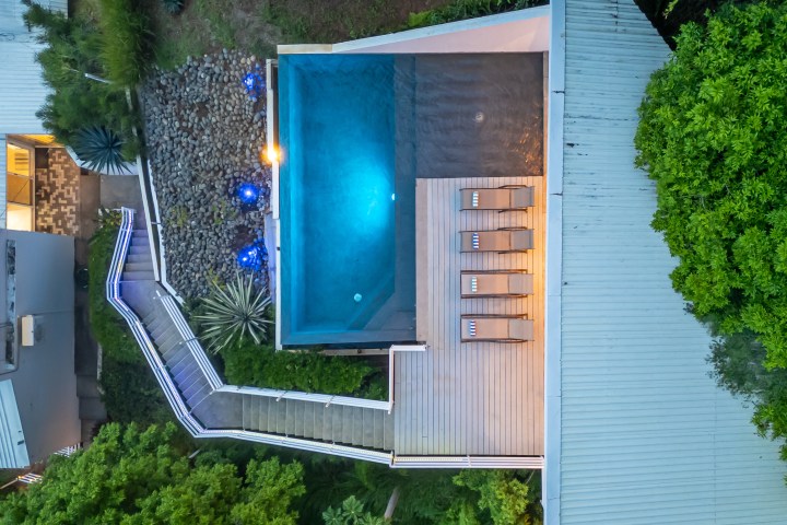 Aerial view of a pool with deck chairs surrounded by trees and pathway.