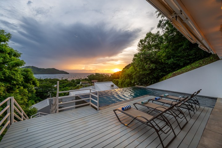 Deck with loungers by a pool overlooking a sunset view of the ocean with boats and lush greenery.