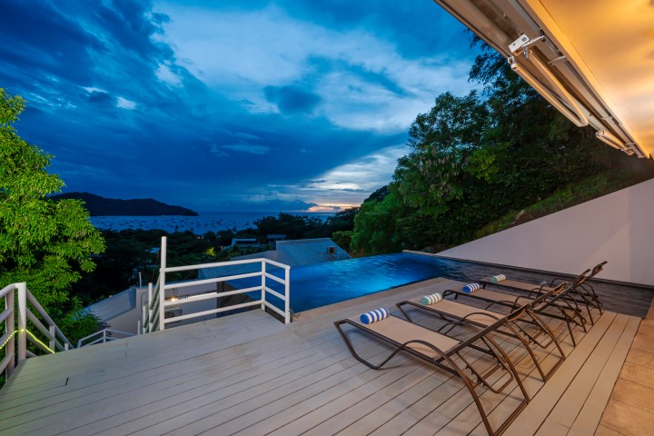 Deck chairs by an infinity pool overlooking a bay at sunset with trees and hills in the background.