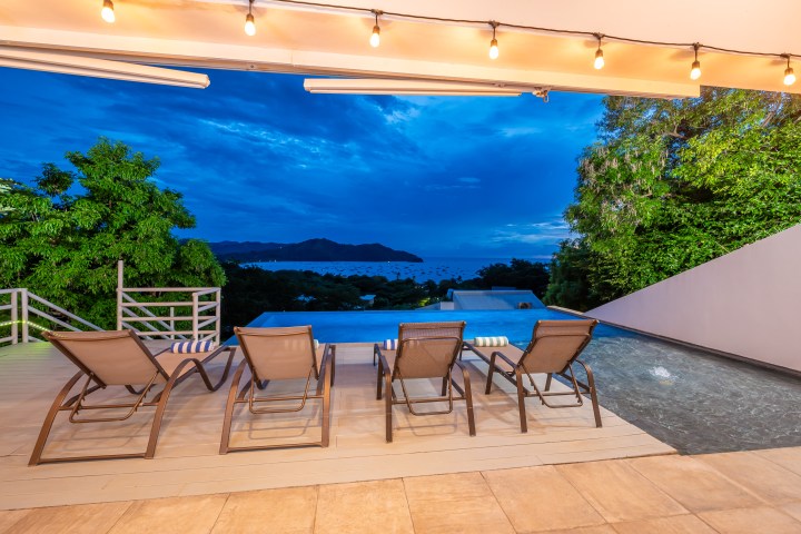 Lounge chairs by an infinity pool with ocean and mountain view at sunset.