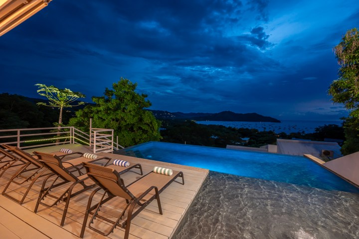 Infinity pool with loungers overlooking ocean and mountains at dusk.