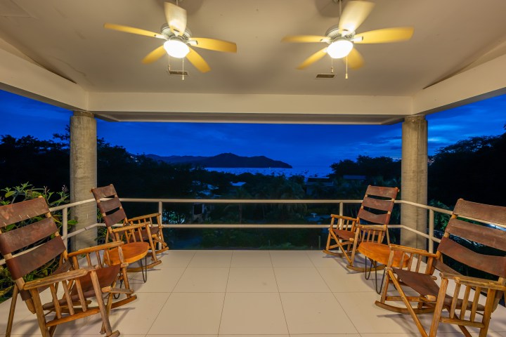 Covered patio with wooden chairs, ceiling fans, and ocean view at dusk.