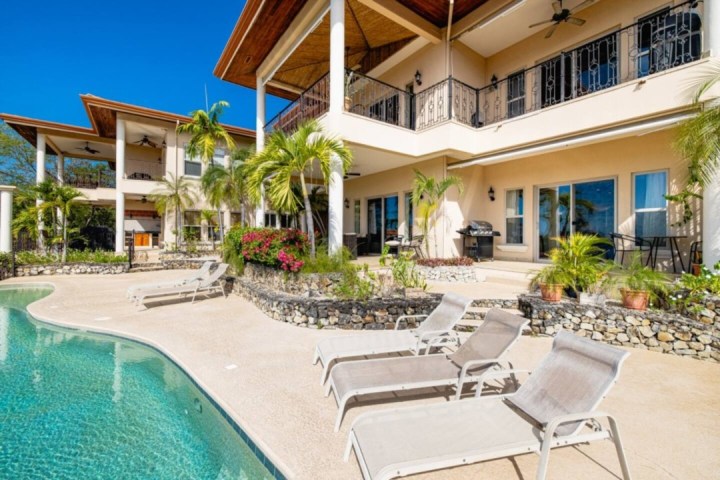 Luxurious villa with pool, sun loungers, palm trees, and multiple balconies under a clear blue sky.