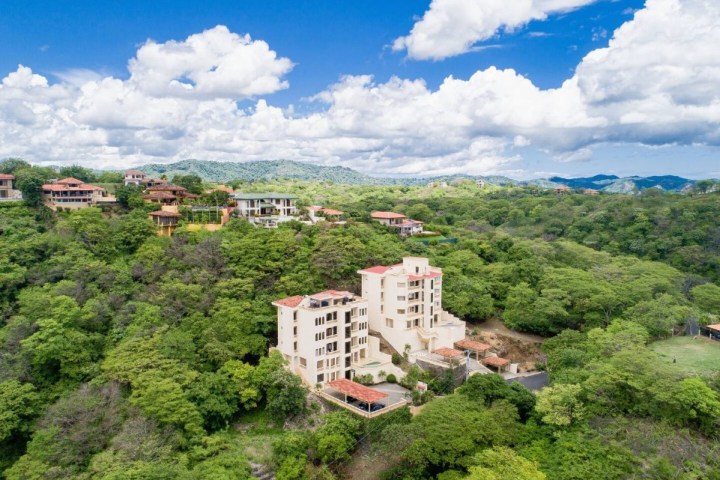 Aerial view of a hillside with buildings surrounded by lush greenery under a blue sky with clouds.