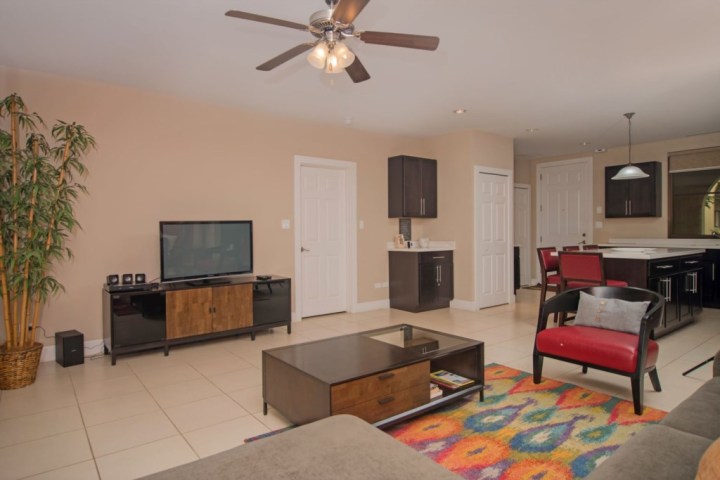 Living room with TV, red chair, coffee table, colorful rug, and ceiling fan; kitchen visible in background.