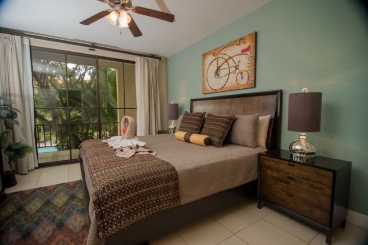 Bedroom with large bed, green wall, bicycle artwork, ceiling fan, and glass doors to a garden view.
