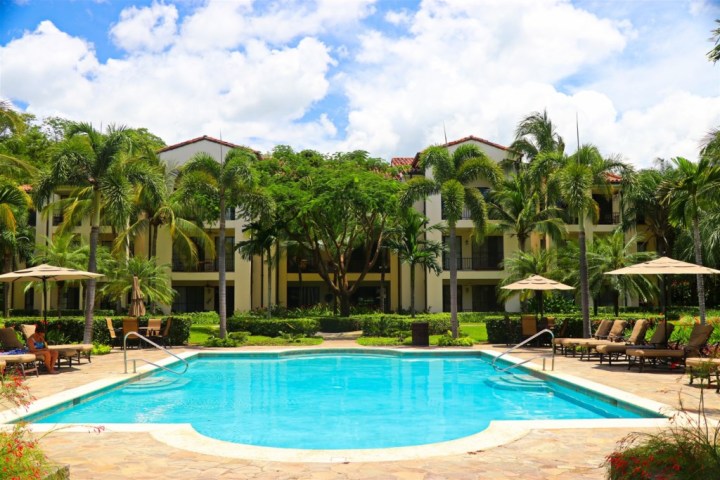 Tropical resort pool with palm trees, sun loungers, and a building in the background under a partly cloudy sky.