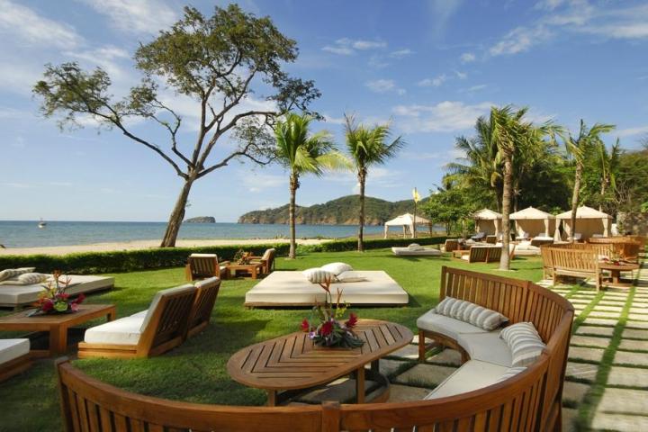 Beachfront patio with wooden furniture, palm trees, and ocean view under a clear blue sky.