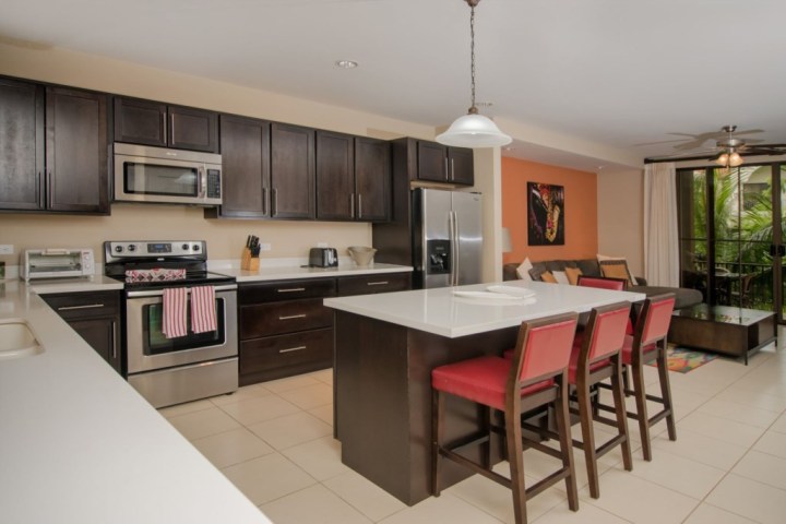 Modern kitchen with dark cabinets, island, red chairs, and stainless steel appliances, open to living area.