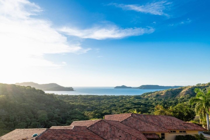 Scenic ocean view over hillside with red-roofed house, blue sky, and tree-lined hills.
