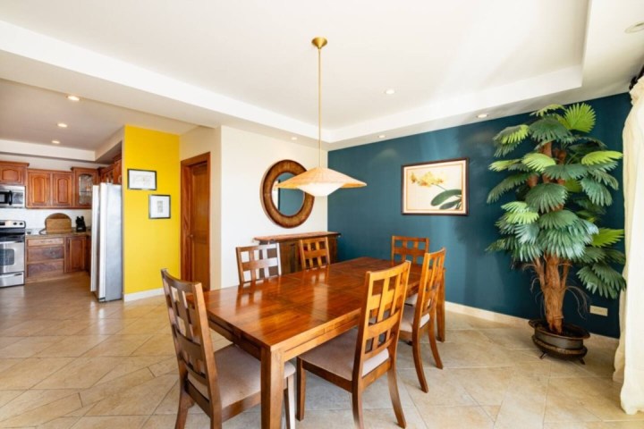 Dining room with wooden table, six chairs, teal accent wall, and large plant beside the kitchen.