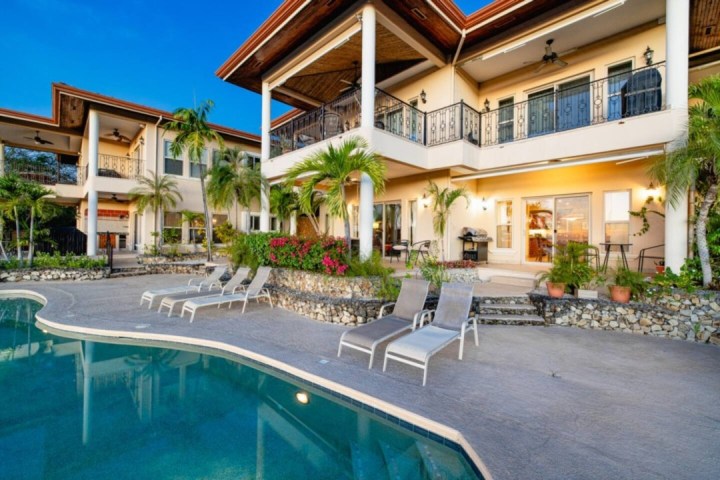 Luxurious two-story house with balconies, poolside lounge chairs, and palm trees around a large swimming pool.
