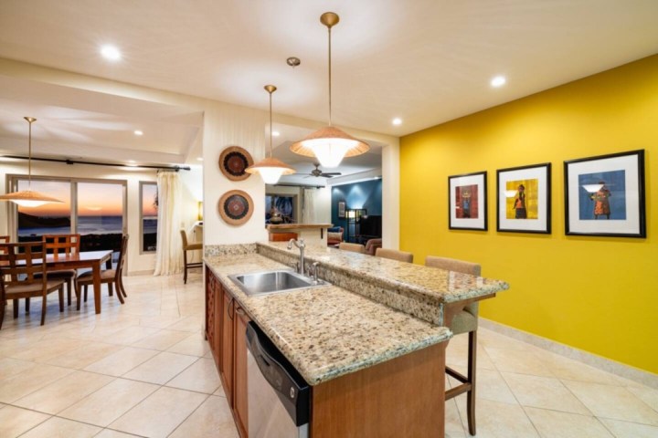 Modern kitchen and dining area with granite island, yellow accent wall, and sunset view.