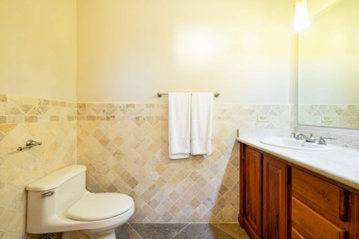 Bathroom with toilet, wooden vanity, and tiled walls. White towel on rack and brightly lit mirror.