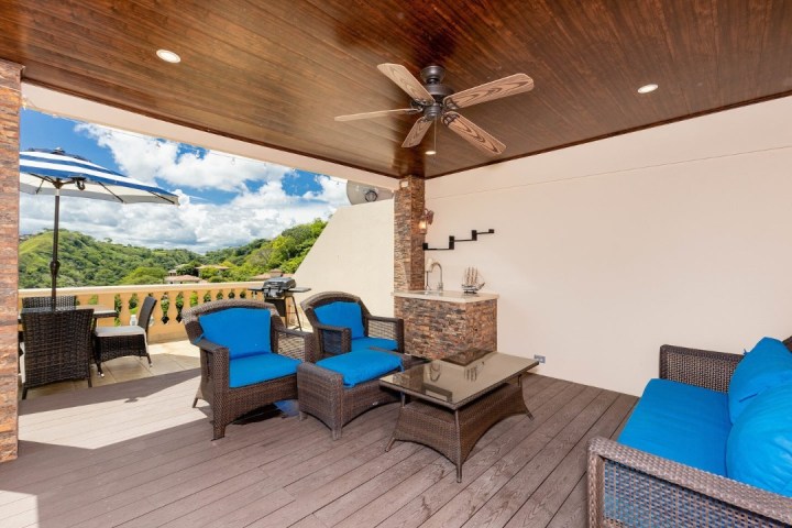 Outdoor patio with wicker furniture, blue cushions, ceiling fan, and a view of hills.