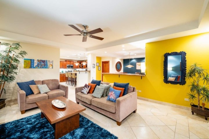 Bright living room with yellow wall, sofas, blue rug, ceiling fan, and view into a kitchen.