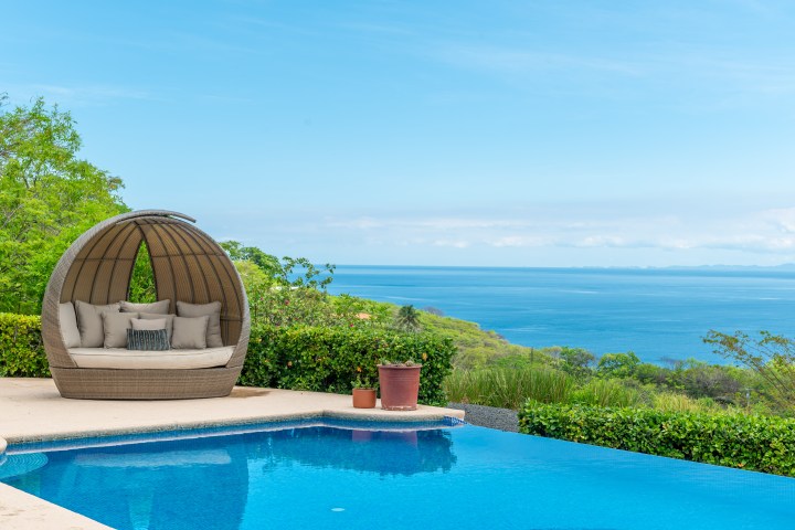 Outdoor lounge by pool with a sea view, featuring a large cushioned wicker chair and potted plants.