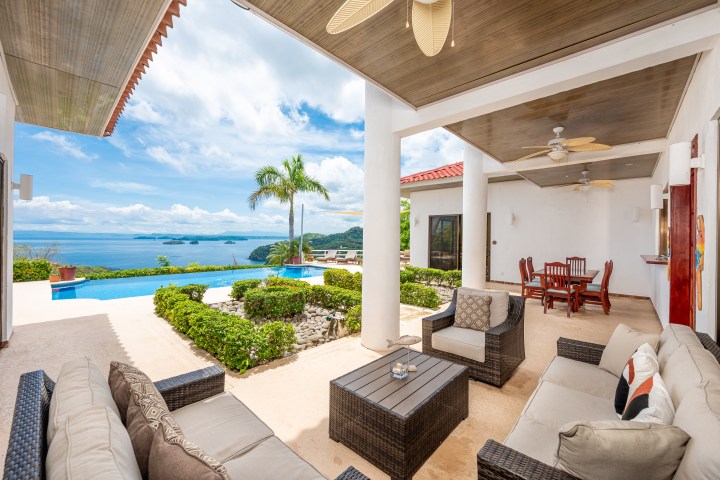 Covered patio with seating, overlooking pool and ocean, with palm trees and blue sky.