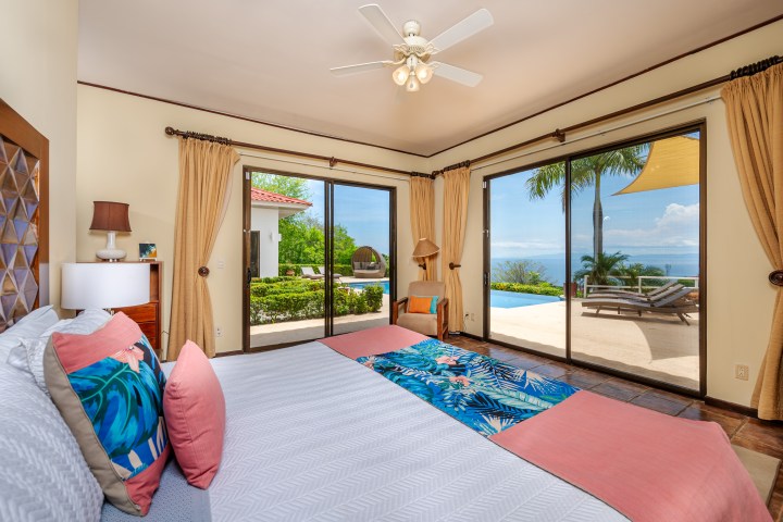Bedroom with ocean view, bed in foreground, glass doors leading to patio with pool and lounge chairs.
