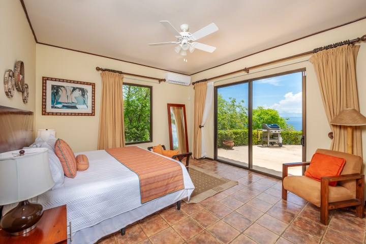Bright bedroom with a large bed, armchair, and sliding doors leading to a patio overlooking the ocean.