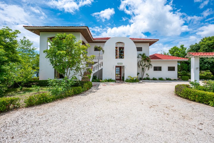 Two-story white house with red roof, surrounded by trees, under a blue sky.