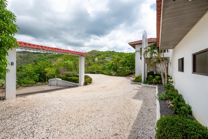 Gravel driveway leading to white house with red-tiled roofs and lush greenery under cloudy sky.