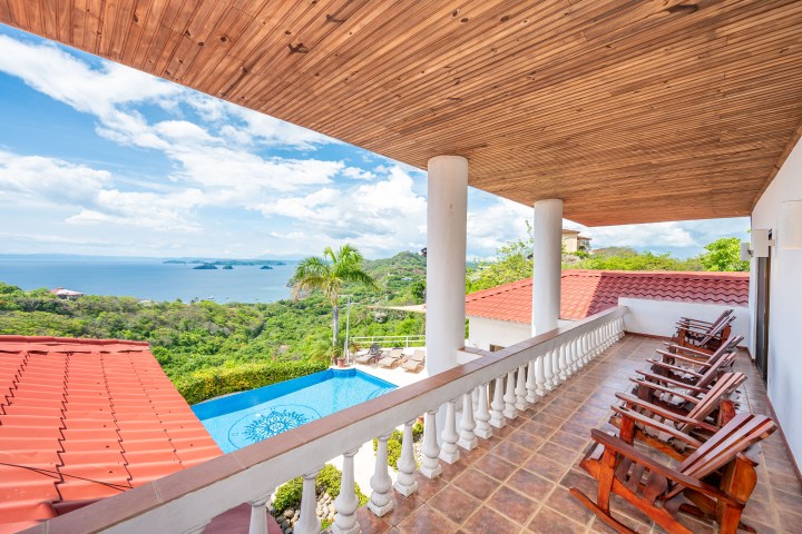 Ocean view from a balcony with wooden chairs, infinity pool, and lush greenery.
