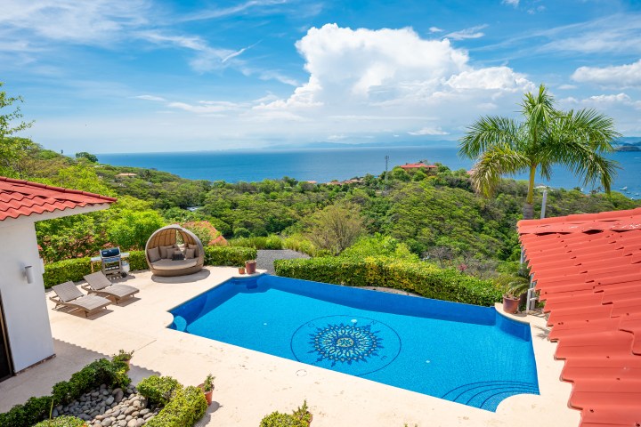 Villa rooftop pool with ocean view, sun loungers, and a daybed surrounded by greenery and palm trees.