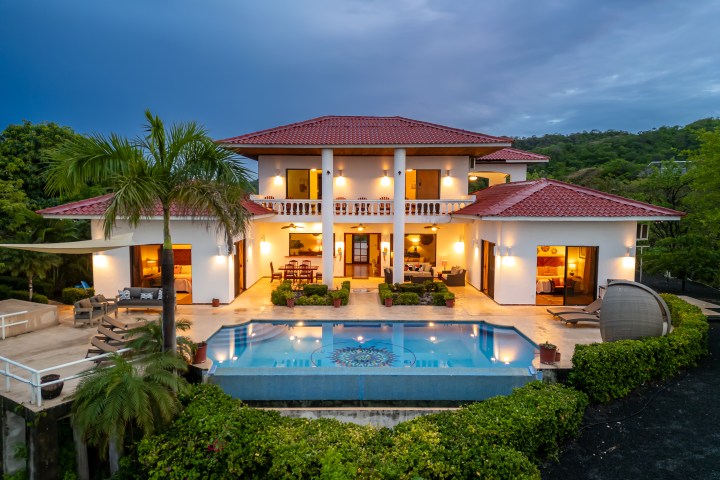 Luxury villa with pool and palm trees at dusk, featuring red roof and lit-up white exterior.