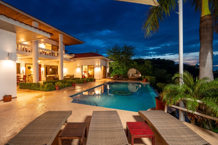 Luxurious villa with pool and lounge chairs at dusk, surrounded by greenery.