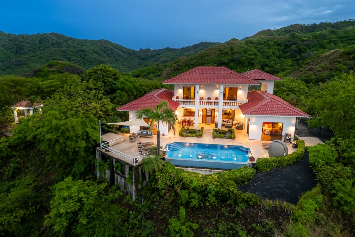 Luxurious villa with red roof and pool, surrounded by lush greenery and hills at dusk.