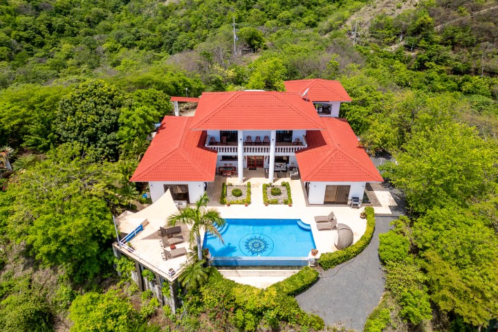 Aerial view of villa with red roofs, pool, and surrounding green foliage.