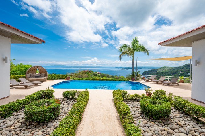 Luxury villa with pool, ocean view, lounge chairs, and palm trees under a blue sky.