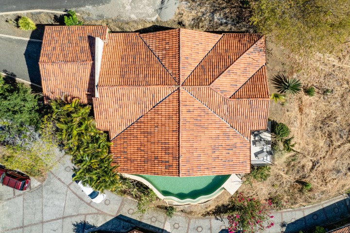 Aerial view of a hexagonal house with a red-tiled roof surrounded by trees and a driveway.