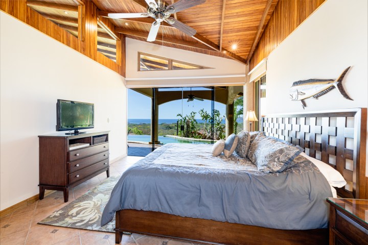 Bedroom with wooden ceiling, bed, TV, and ocean view through large windows.