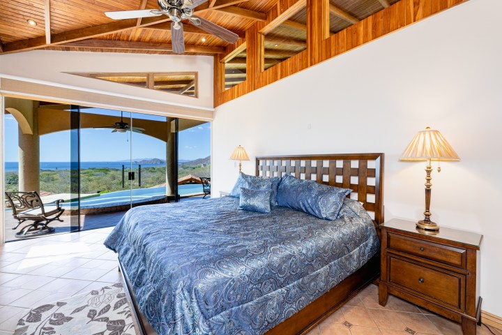 Spacious bedroom with large bed, ocean view through glass doors, and wooden ceiling.