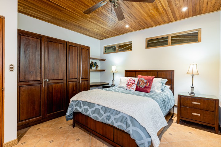 Cozy bedroom with wooden ceiling, large wardrobe, bed with fish-themed pillows, and bedside lamps.