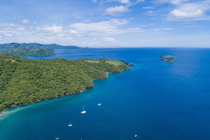 Aerial view of a coastal landscape with lush green vegetation, ocean, and a few boats.