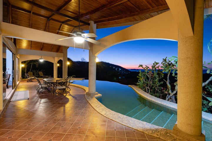 Covered patio with dining set, ceiling fan, and pool overlooking a scenic sunset view.