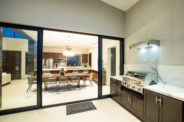 Modern indoor kitchen with large dining table, visible through glass doors, next to an outdoor grill area.