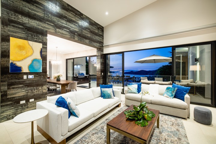 Modern living room with white sofas, blue cushions, large glass doors, and view of a terrace with outdoor seating.
