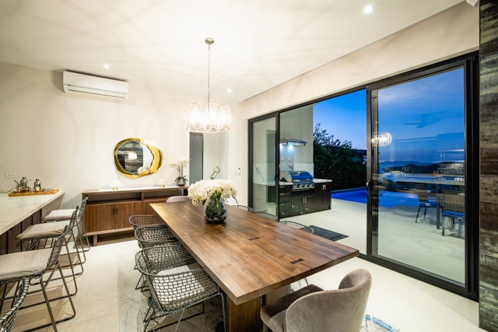 Modern dining room with wooden table, wire chairs, and open sliding doors to a patio and pool at dusk.