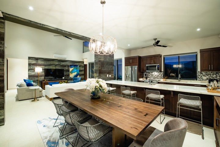 Modern kitchen and living room with wooden table, counter stools, and a large chandelier.