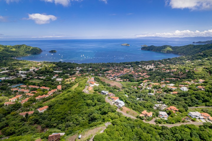 Aerial view of a coastal town with lush greenery, red-roofed buildings, and a bay with boats under a blue sky.