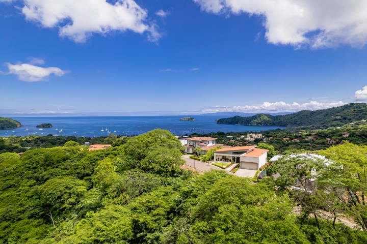Aerial view of a coastline with houses, lush greenery, and boats on the ocean under a blue sky with clouds.